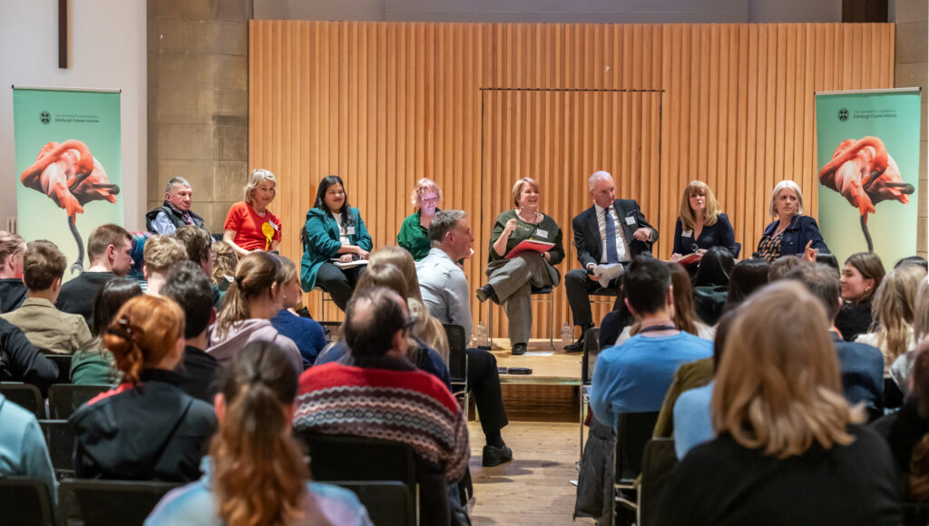 A panel of seven people sits on a stage in front of an audience, engaged in a discussion. The room has wooden panelling and two banners with a red and black graphic on either side of the stage.