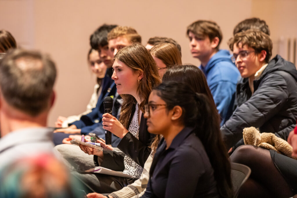 A young woman in a crowd speaks into a microphone during an indoor event, surrounded by other attentive attendees seated in rows.