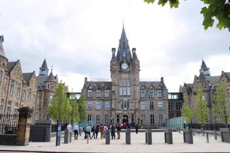 A large stone building with a central clock tower, arched windows, and people walking and gathering out front, surrounded by trees and modern paving. The sky is cloudy.