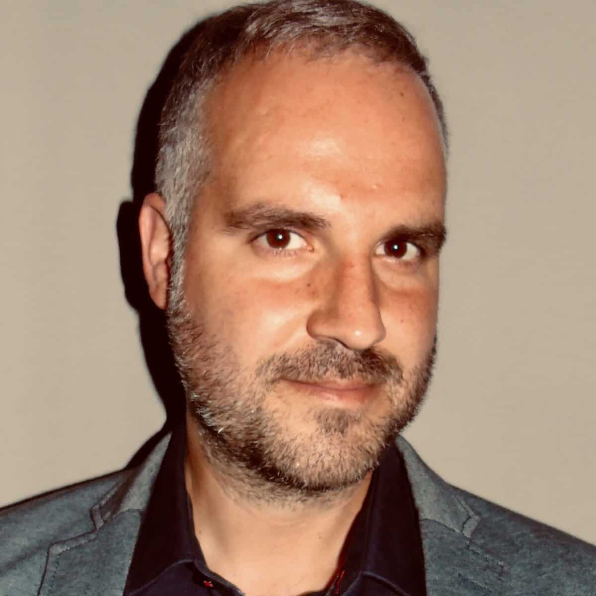 A man with short grey hair and a trimmed beard is wearing a dark collared shirt and a grey blazer, standing in front of a plain, light-coloured background and looking at the camera with a slight smile.