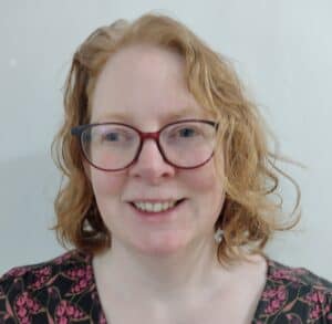 A woman with wavy, shoulder-length red hair and glasses is smiling. She is wearing a dark top with a pink floral pattern and is standing in front of a plain white background.