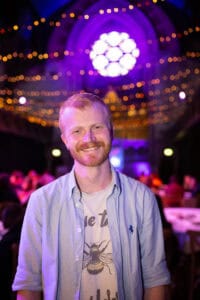 A smiling man with blond hair and a beard stands in a warmly lit venue with fairy lights overhead and a stained glass window in the background. He wears a light shirt over a graphic t-shirt.