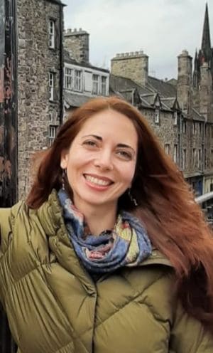 A woman with long reddish-brown hair, wearing a green jacket and patterned scarf, smiles outdoors in front of old stone buildings with tall chimneys and gabled roofs.