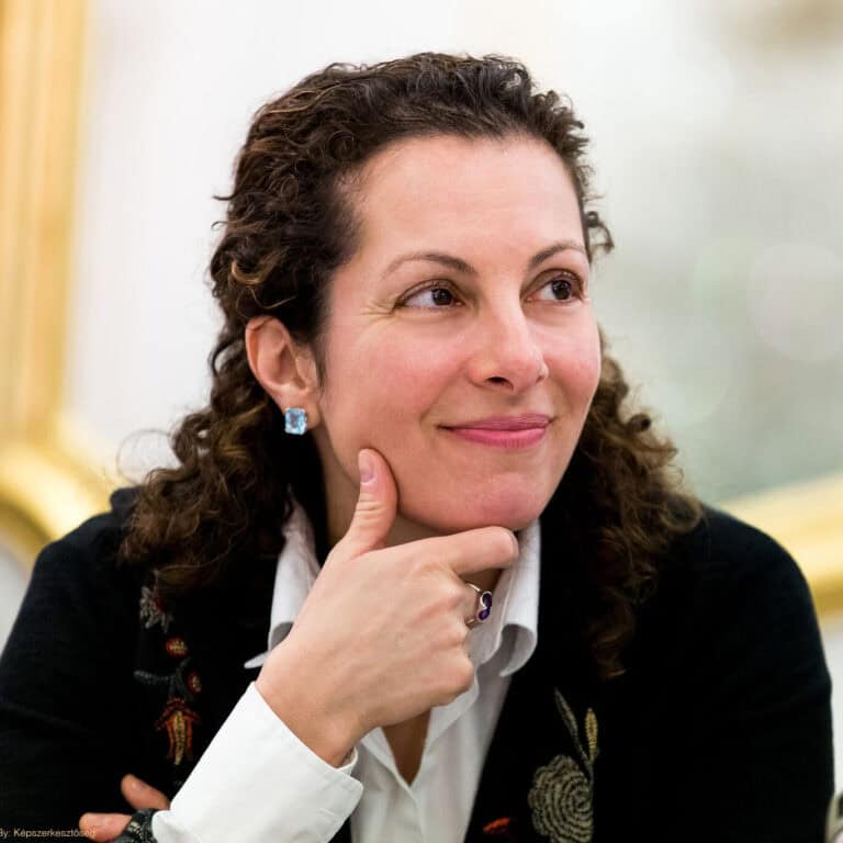A woman with curly brown hair, wearing a white collared shirt and a dark jacket, smiles softly whilst resting her chin on her hand. She wears a square blue earring and a purple ring. The background is softly blurred.