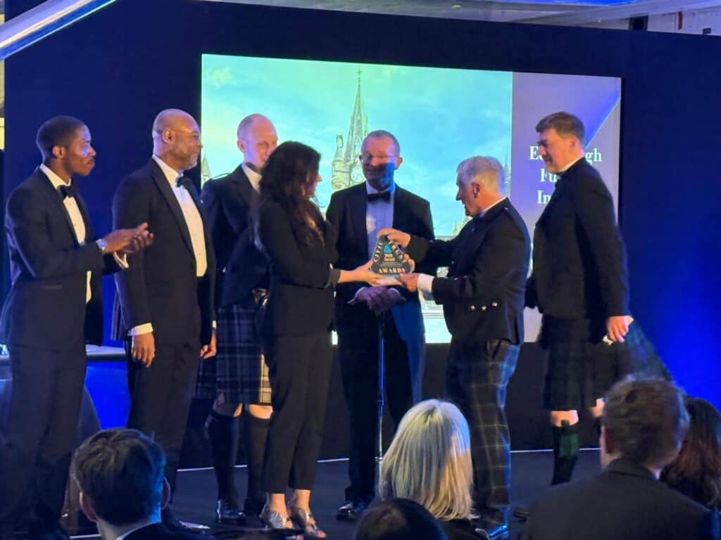 A group of people dressed in formal attire, some in kilts, stand on stage as a woman presents an award to a man. Others look on and applaud. A projection screen is visible in the background.