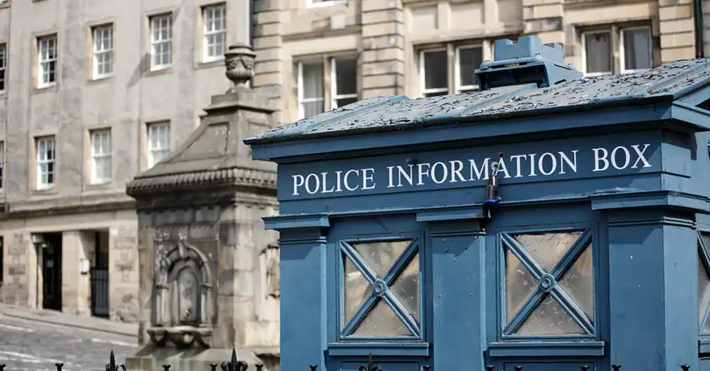 A blue police information box with white lettering stands in front of historic stone buildings on a city street. The scene appears quiet and the architecture is classic and traditional.