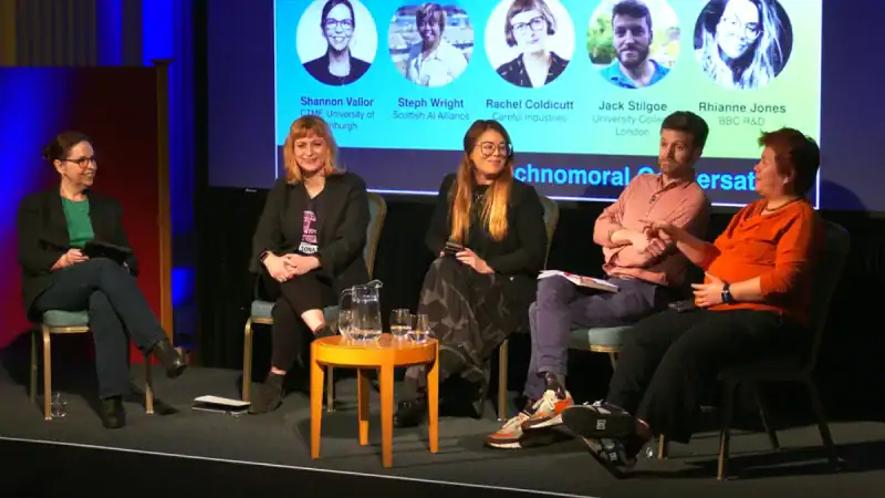 Five people sit on a stage, engaged in a panel discussion. A large screen behind them displays headshots, names, and affiliations of the speakers. The group appears attentive, with one person speaking whilst others listen.
