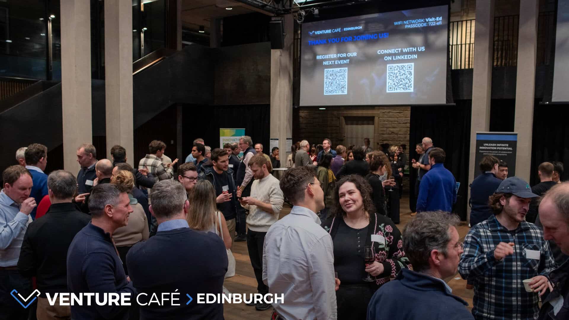 A large group of people socialise and network at an indoor event, with two big screens displaying QR codes and Venture Café Edinburgh branding in the background.