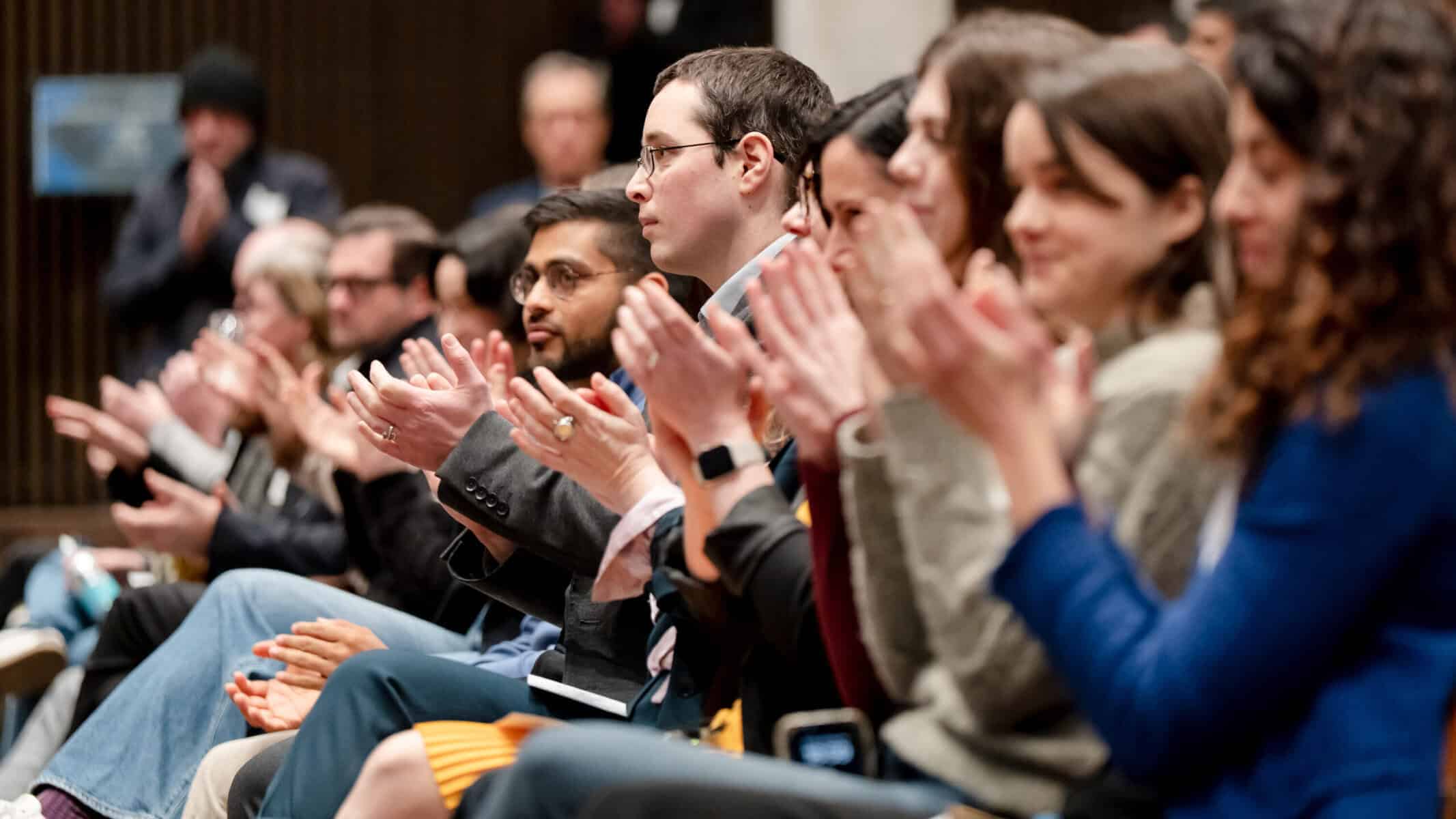 A group of people sitting in rows and applauding, focused on something happening out of view. The attendees appear engaged and attentive in a formal indoor setting.