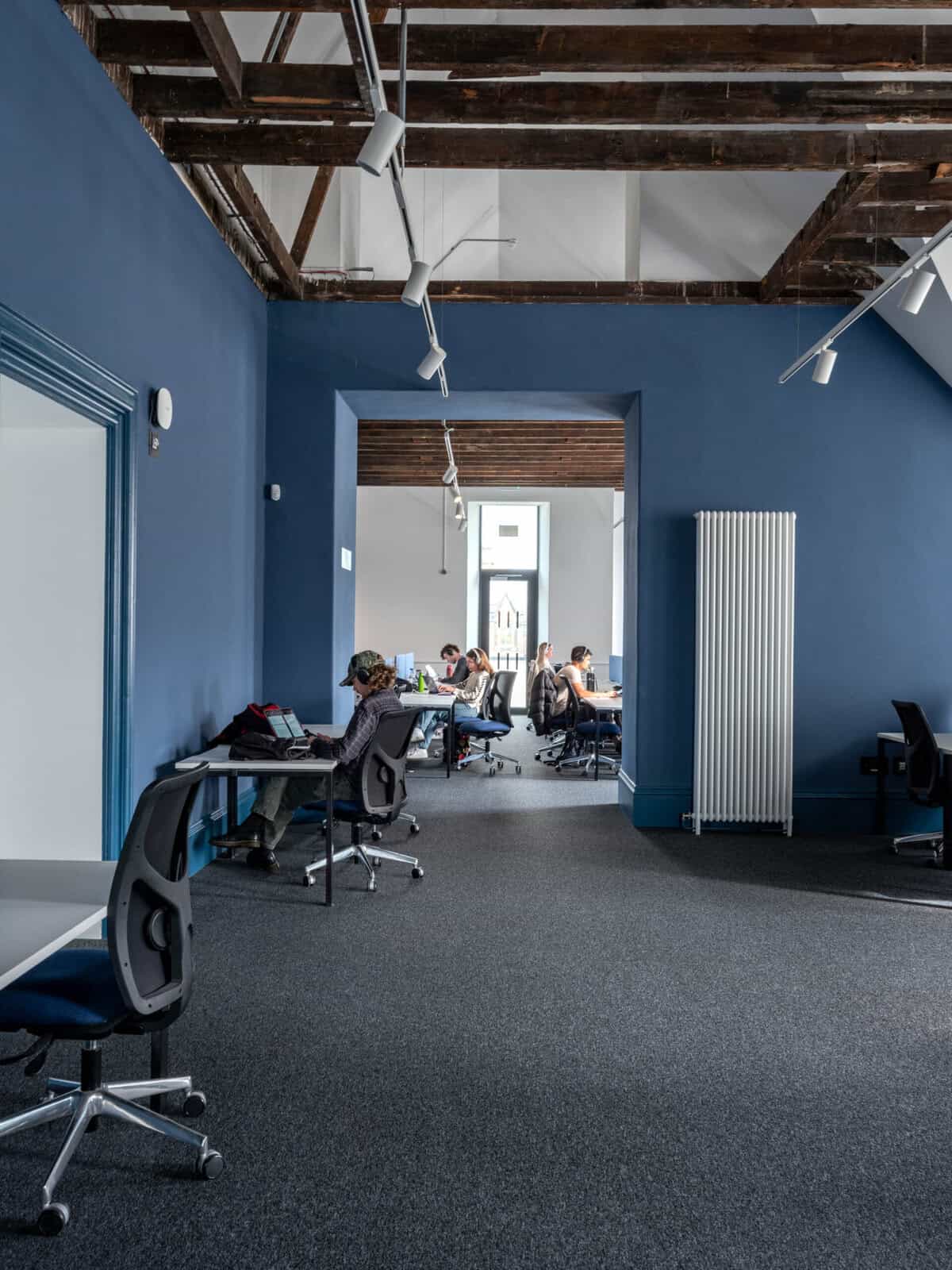 People work at desks in a modern space with blue walls, exposed wooden beams, and large windows. The room features ergonomic chairs, a radiator, and ample natural light.