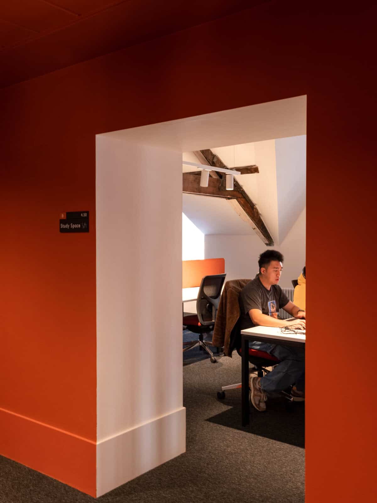 A person sits at a desk with a laptop in a cosy study space with orange walls and exposed wooden beams, visible through a doorway. A sign beside the door reads "Study Space.