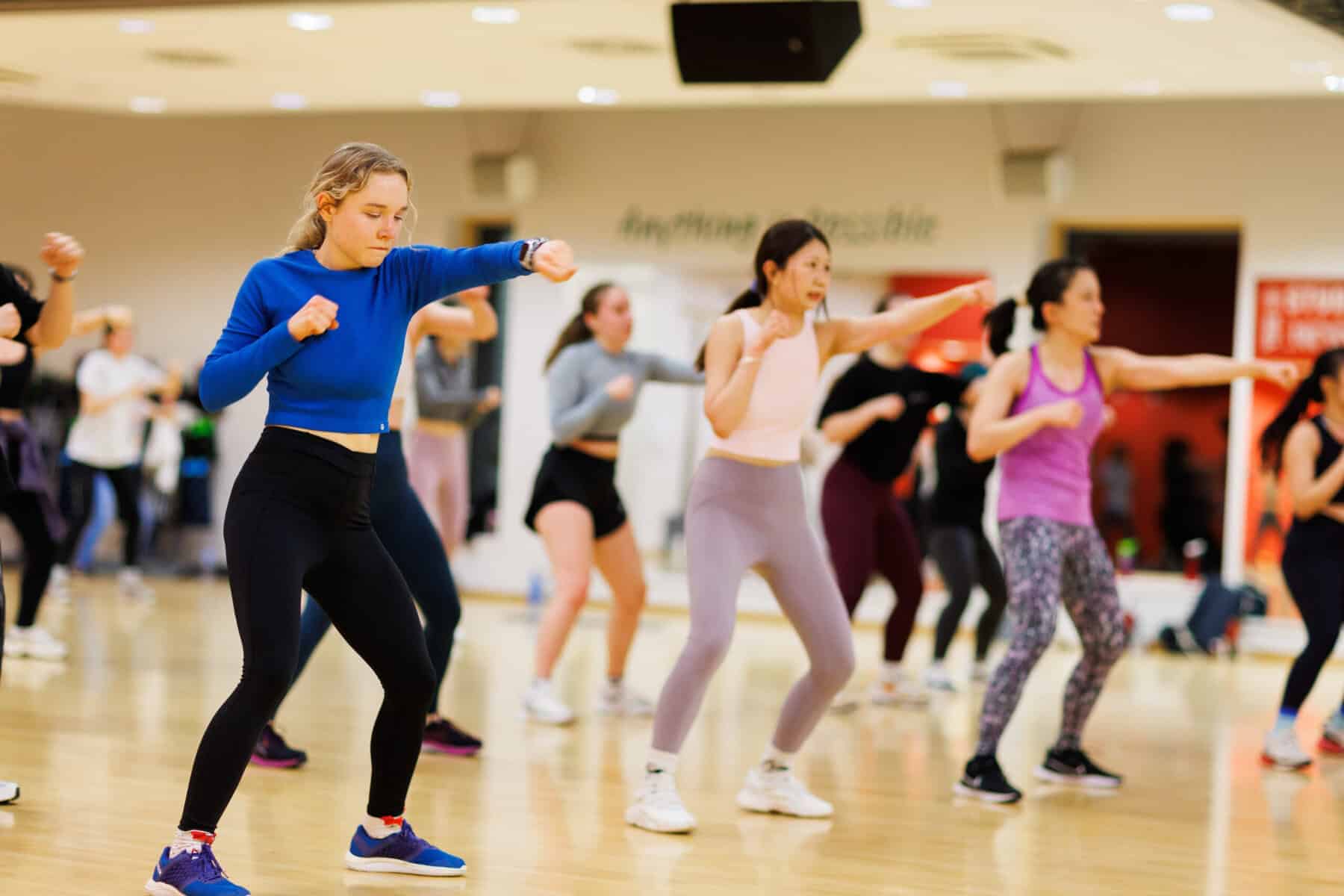 A group of people in sportswear participate in a fitness class in a gym, punching the air and moving in unison. The focus is on a woman in a blue top and black leggings at the front.