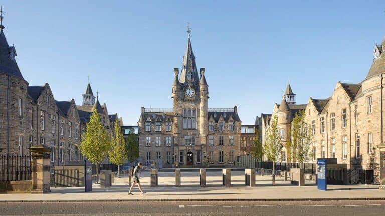 A person walks past a historic, Gothic-style stone building with a central clock tower, surrounded by trees and blue sky on a sunny day.