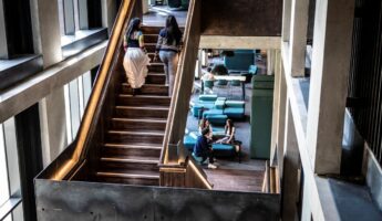 Two people walk up a wooden staircase inside a modern building with large windows, whilst four others sit and talk on teal chairs and sofas in the lounge area below.