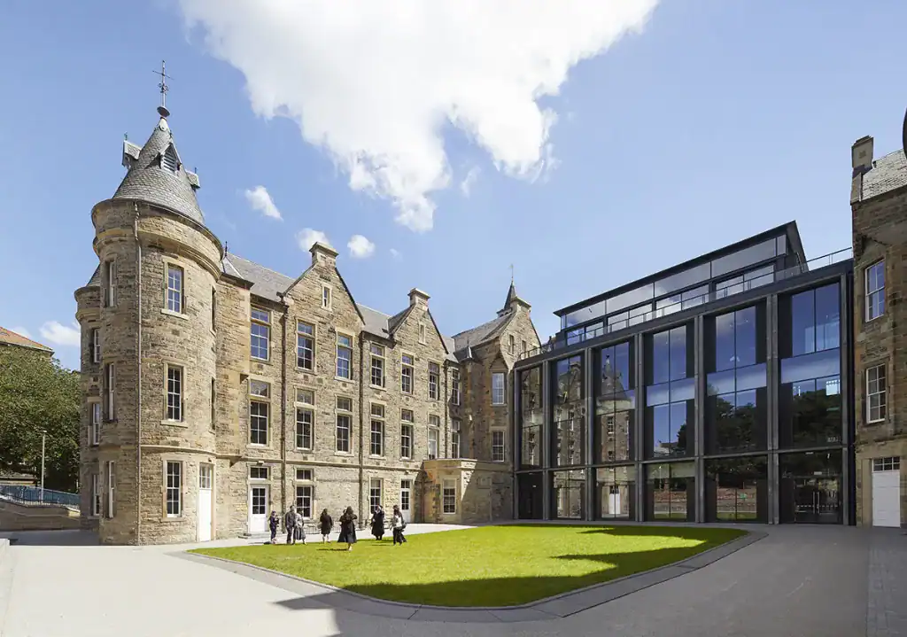 Historic stone building with round towers and pointed roofs on the left, connected to a modern glass extension on the right, surrounding a grassy courtyard with several people walking. Blue sky with scattered clouds above.