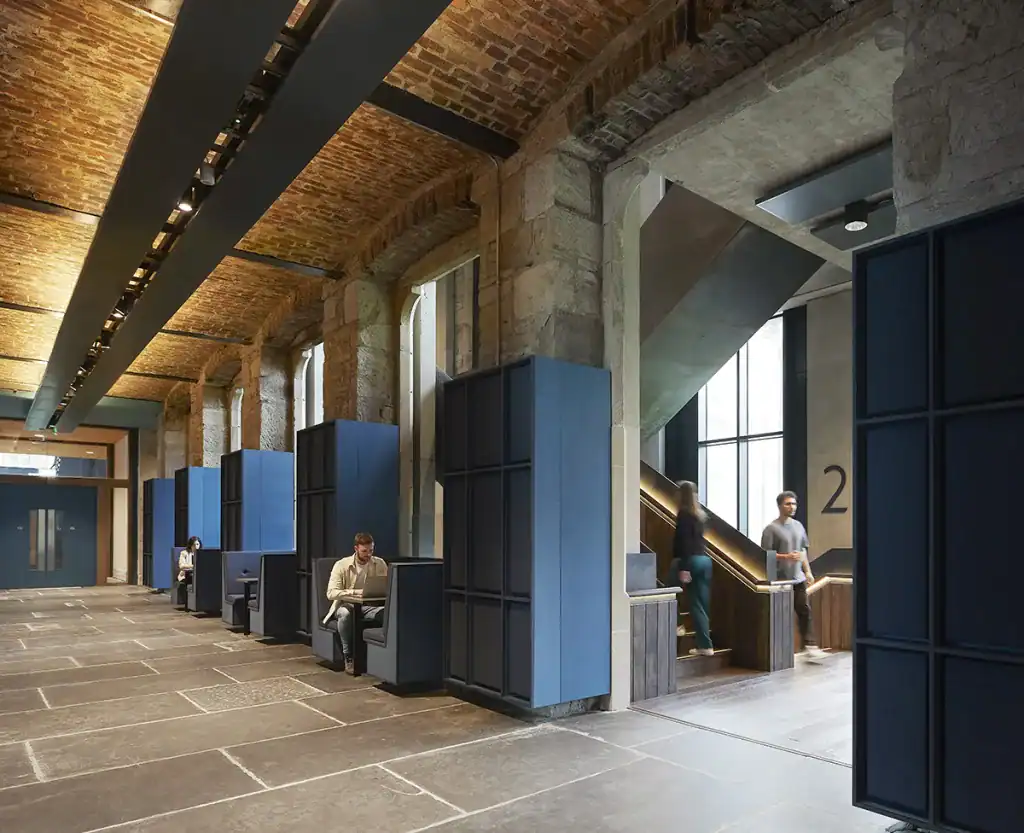 A spacious, modern interior with exposed brick ceilings, stone walls, and blue seating booths along one side. People are seated and walking near a staircase with large windows, creating an open and airy atmosphere.