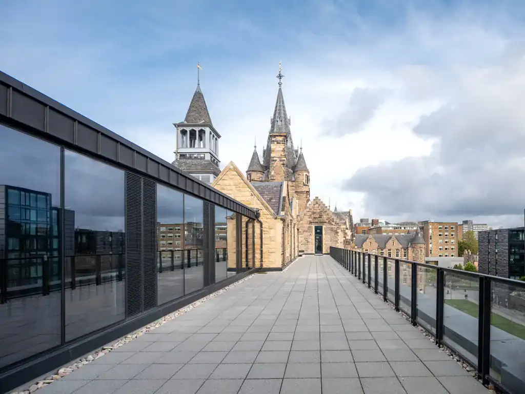 A paved rooftop terrace with black railings overlooks modern and historic stone buildings, including a church with tall spires, under a partly cloudy sky.