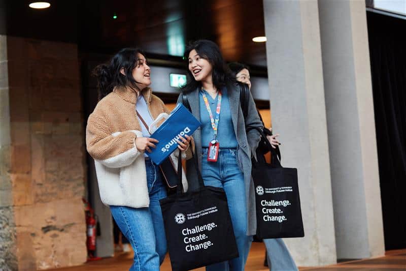 Three young peoplewearing casual clothes smile and talk whilst walking indoors, carrying black tote bags that read "Challenge. Create. Change." 
