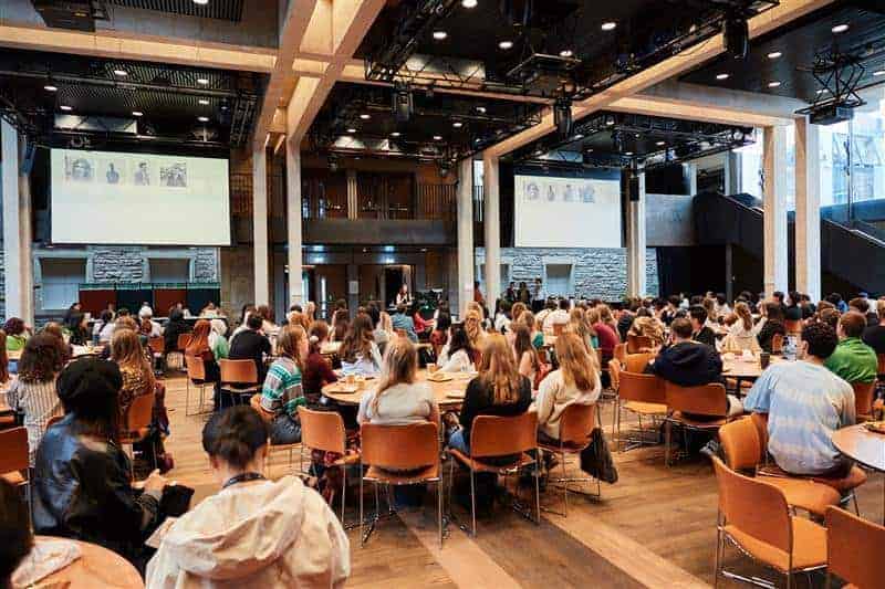 A large group of people seated at round tables in a spacious modern conference room, facing a stage with speakers and two large projection screens displaying images.