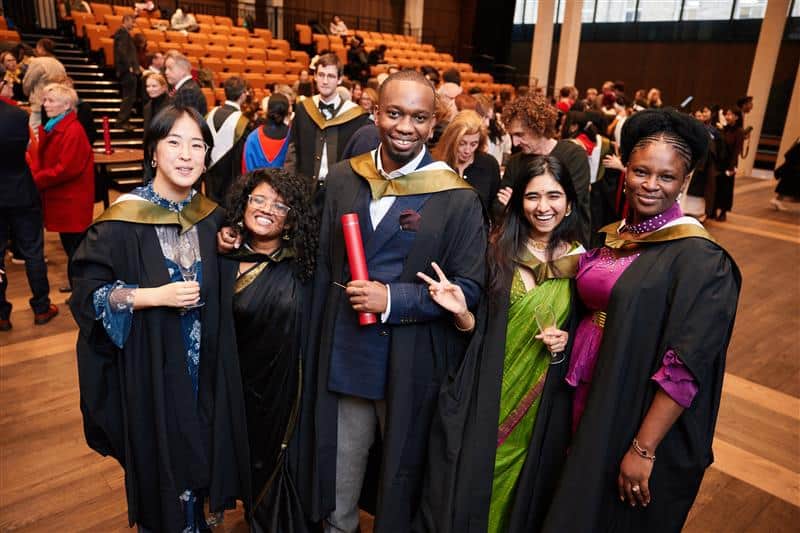 Five graduates in caps and gowns smile and pose together in a hall, with one holding a red degree certificate. Other people and empty seats are visible in the background.