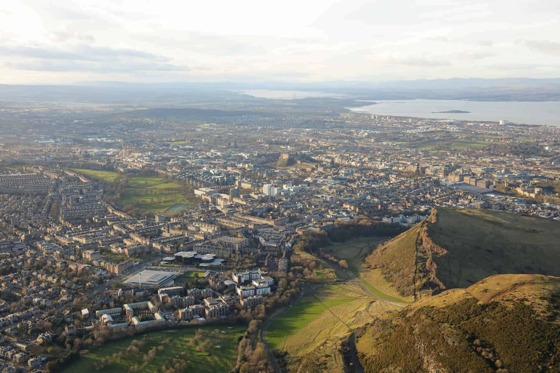 Aerial view of a cityscape with densely packed buildings, green parks, and hills in the foreground, leading to a distant coastline under a cloudy sky.