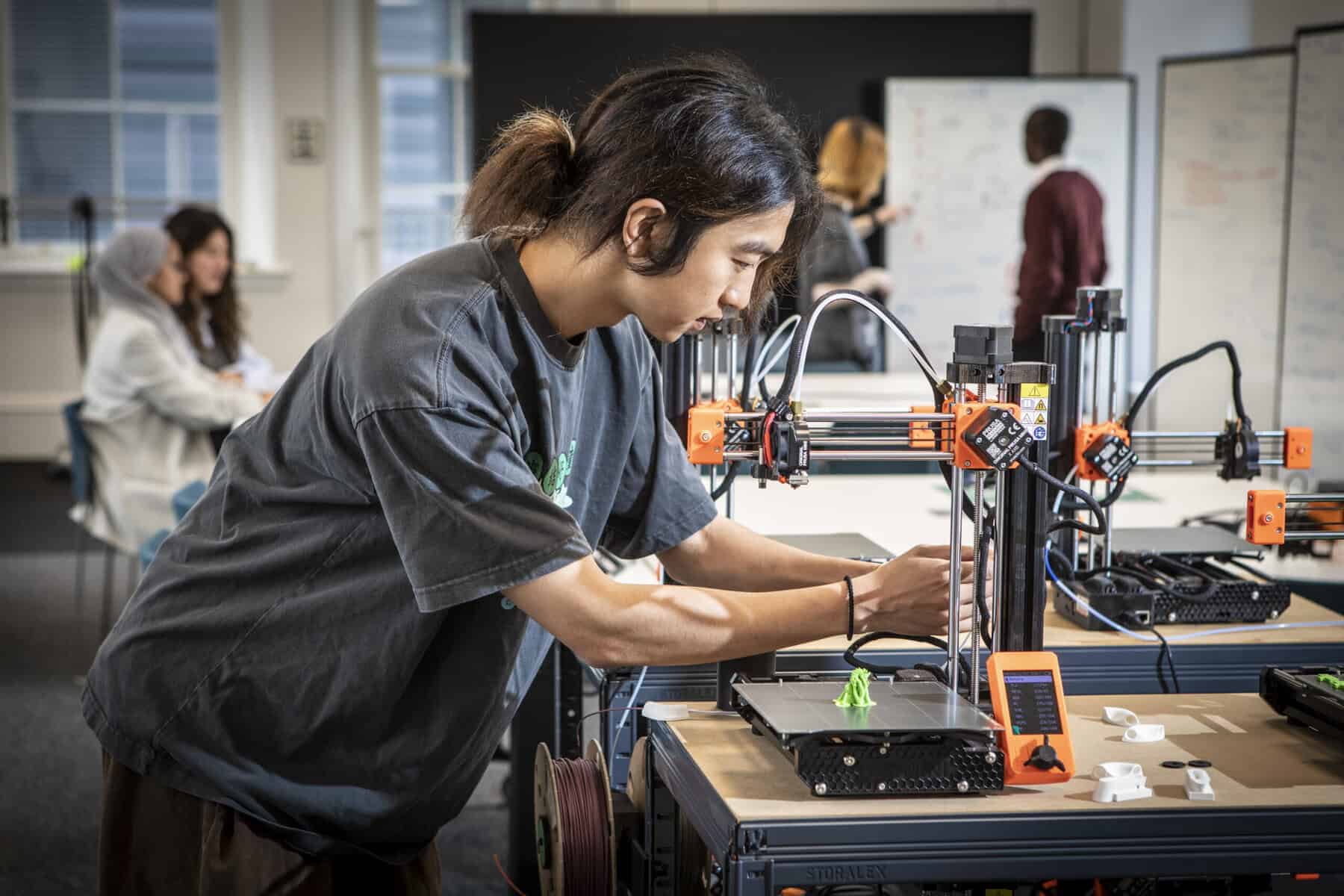 A person adjusts a 3D printer in a classroom or lab setting, with a green object being printed. Other people are working and talking in the background.