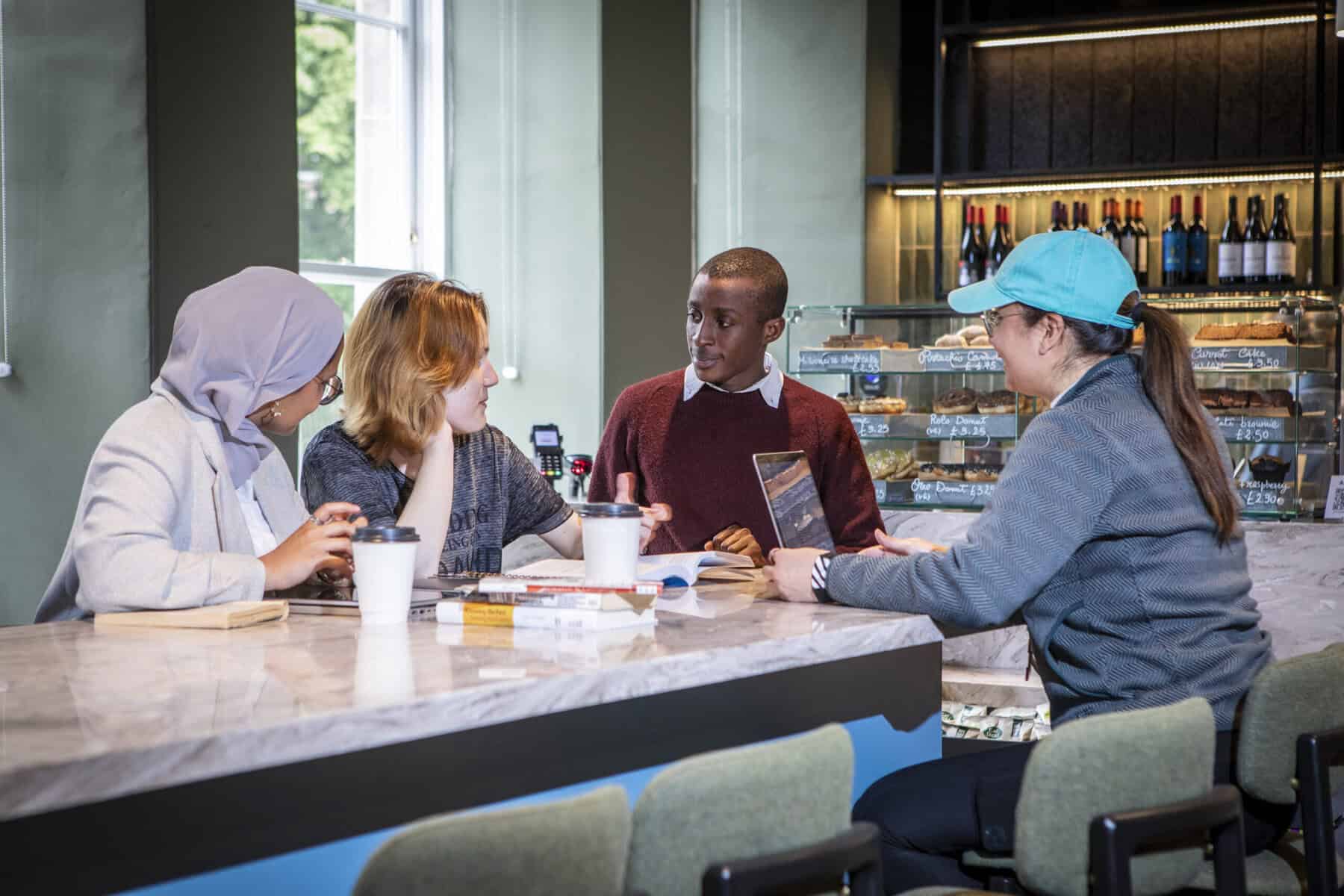 Four people sit around a marble table in a café, engaged in conversation with books, notebooks, and coffee cups. Shelves with pastries and wine are visible in the background.