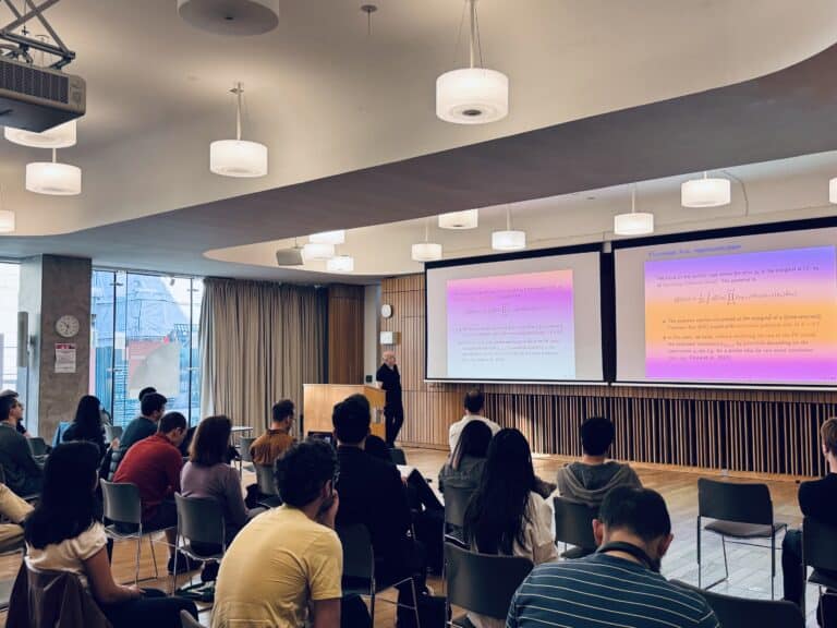 A group of people sit facing a presenter in a modern lecture theatre with large windows. The presenter stands beside two projection screens displaying mathematical formulae and text.