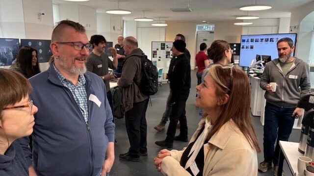 A group of people engaged in conversation at an indoor networking event, with name badges visible. The background shows informational displays, a screen, and others mingling in a well-lit room.
