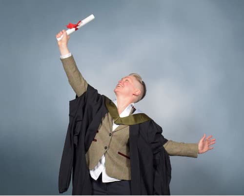 A graduate in a cap and gown holds up a degree triumphantly, smiling and looking upwards, with arms outstretched against a plain blue-grey background.