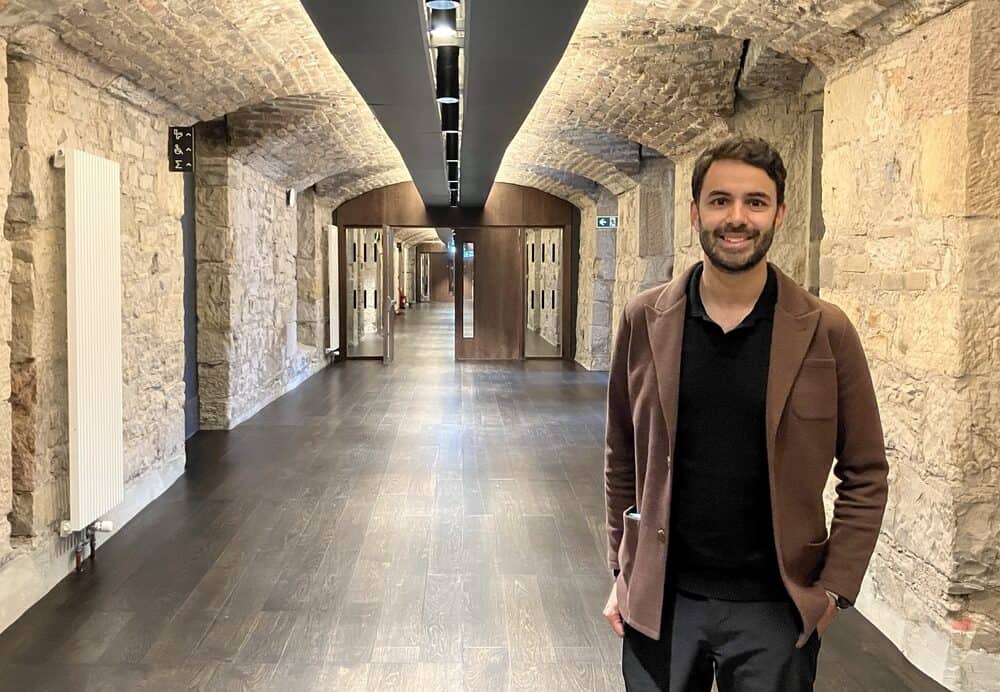 A man with dark hair and a beard, wearing a brown jacket and black shirt, stands smiling in a corridor with stone walls, arched ceilings, and wooden floors. Glass doors are visible at the end of the corridor.