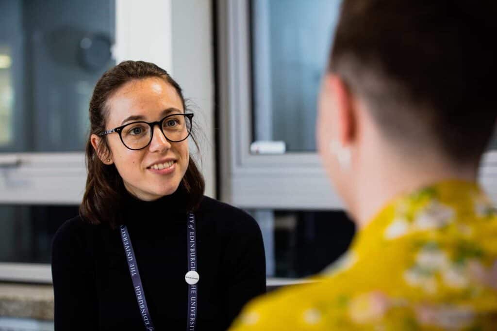A person with glasses and a lanyard smiles while talking to another person in a yellow floral shirt. They are indoors, sitting near a window.