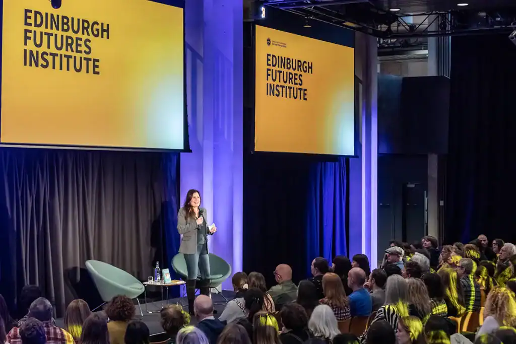 A speaker stands on stage addressing a seated audience at the Edinburgh Futures Institute, with large yellow screens displaying the event name behind her.