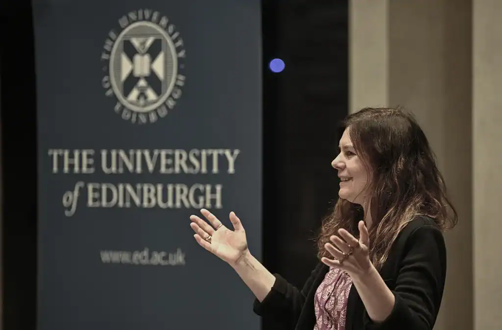 A woman gestures with open hands whilst speaking next to a blue banner that reads "The University of Edinburgh" and displays the university’s crest and website URL.