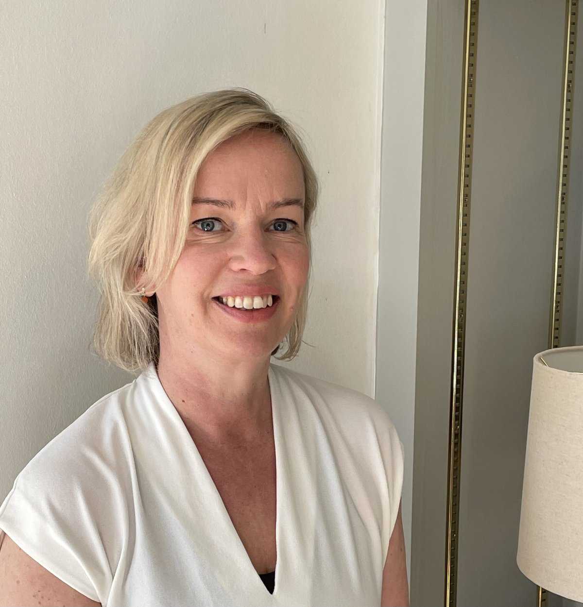 A woman with short blonde hair and a white blouse smiles whilst standing indoors near a white wall and a tall lamp.