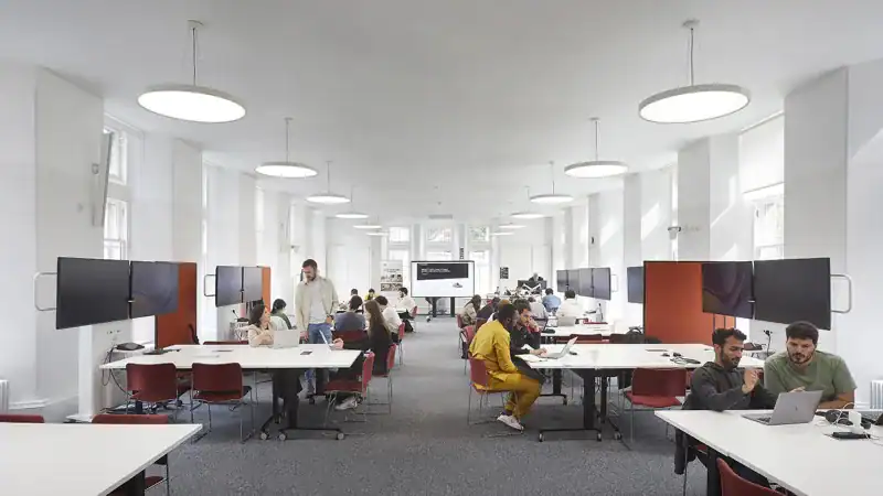 A modern, bright office space with several groups of people working at large white tables, computers and monitors on desks, and large round ceiling lights illuminating the room.