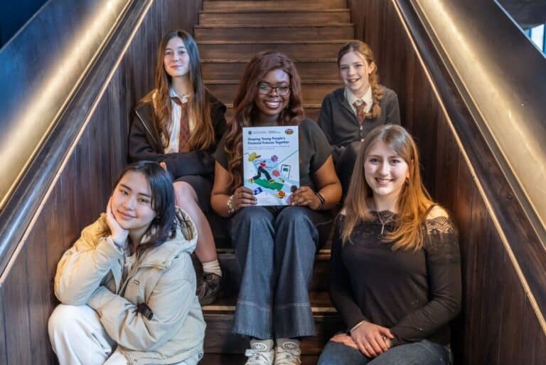 Five young women sit on wooden stairs, smiling at the camera. The woman in the centre holds a colourful booklet. Two women wear school uniforms, while the others wear casual clothes. The setting appears to be indoors.