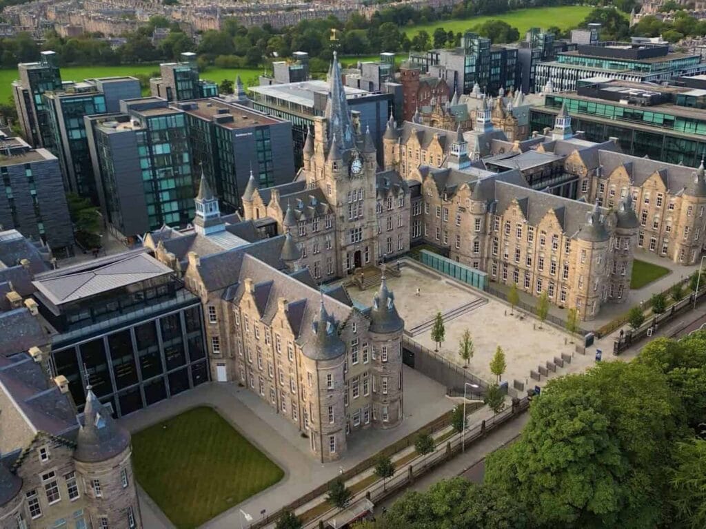 A large historic stone building with pointed towers and a courtyard, surrounded by modern glass buildings and green spaces in an urban setting, viewed from above.