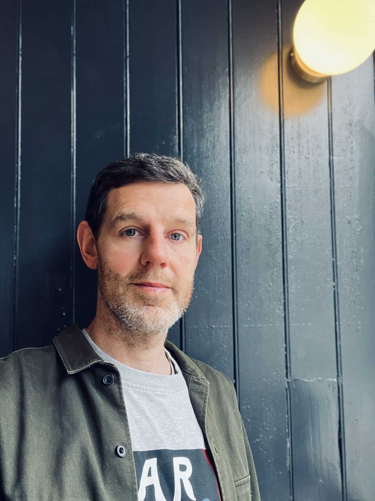 A man with short brown hair and beard stands in front of a dark wooden wall with a round light fitting above him. He is wearing a green jacket over a grey T-shirt.