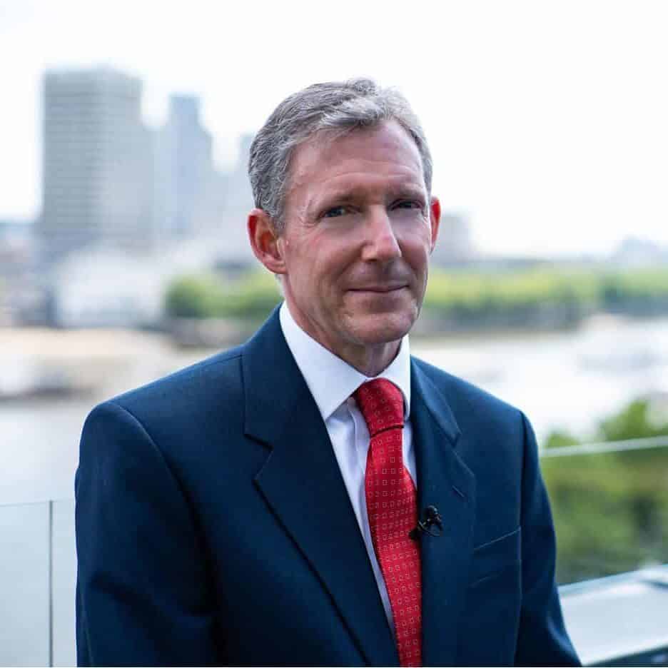 A man in a blue suit and red tie stands on a balcony overlooking a blurred cityscape with tall buildings and a river in the background on a bright day.