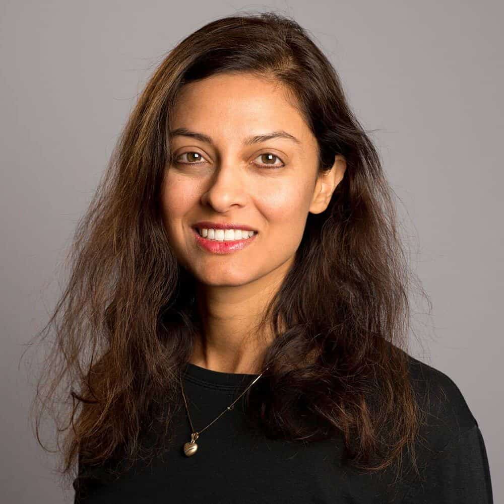 A woman with long brown hair, wearing a black top and a gold necklace, smiles softly at the camera against a plain grey background.
