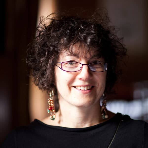 A woman with curly dark hair, wearing glasses, a black top, and dangly earrings, smiles warmly in soft, natural lighting with a blurred background.