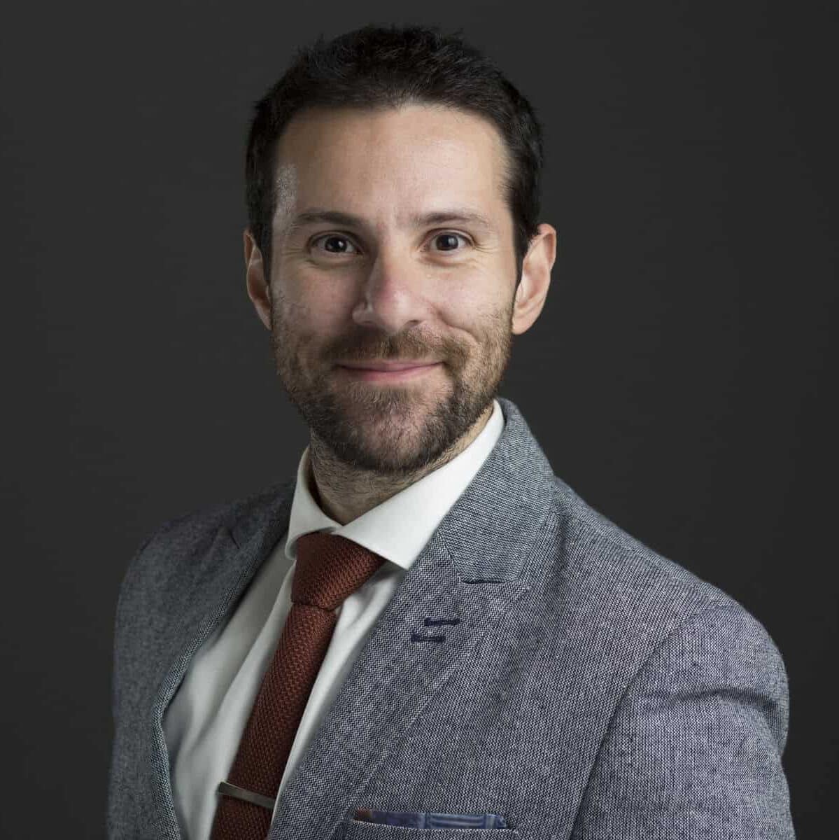 A man with short dark hair and a beard smiles at the camera. He is wearing a grey suit jacket, white shirt, and a red tie, standing against a dark background.