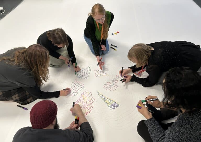 Six people sit in a circle on the floor, collaboratively drawing colourful geometric patterns with markers on a large white sheet. The design radiates from the centre, and everyone is focused on their artwork.