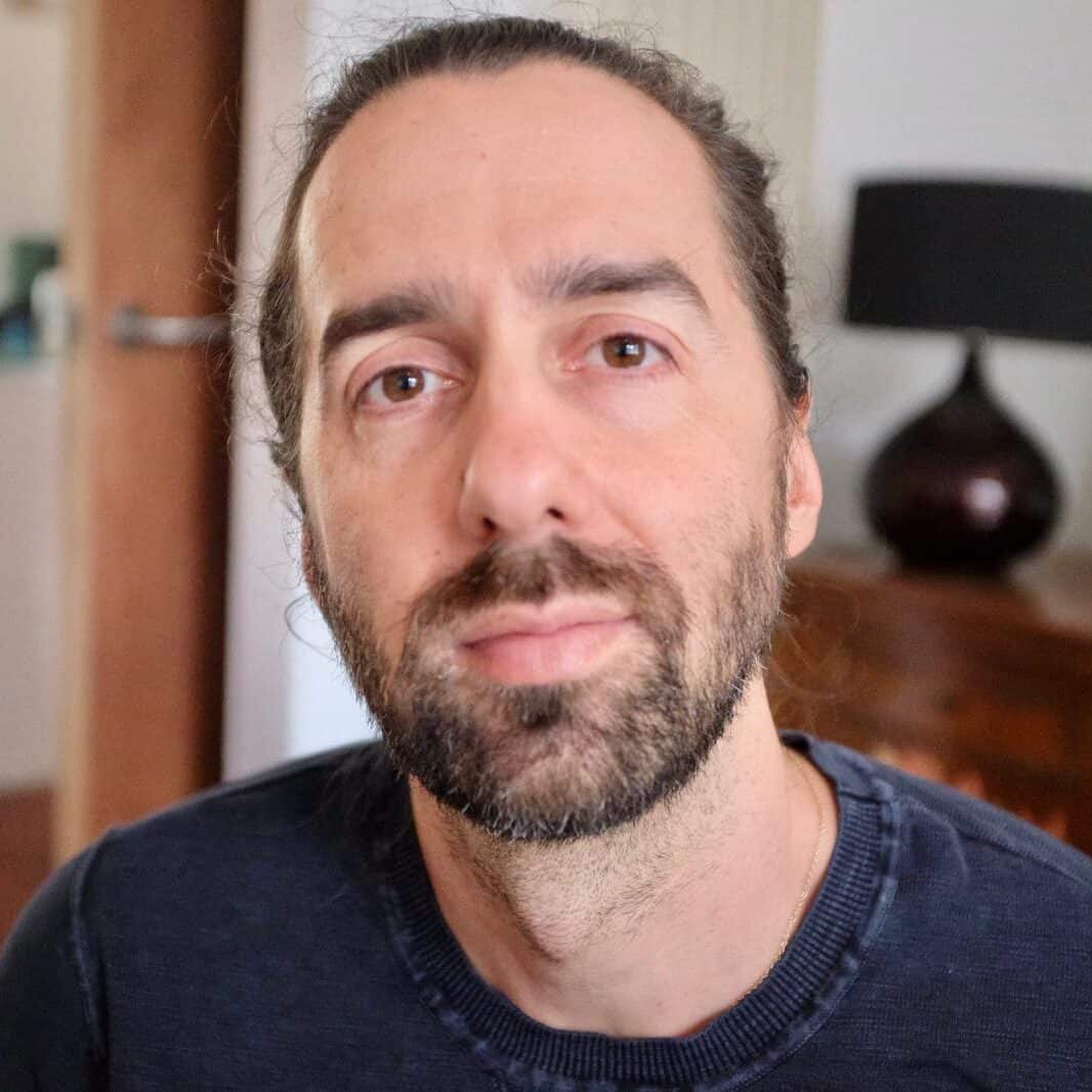 A man with long hair tied back and a beard looks directly at the camera. He is wearing a dark jumper and is indoors, with a lamp, framed photos, and wooden furniture in the background.