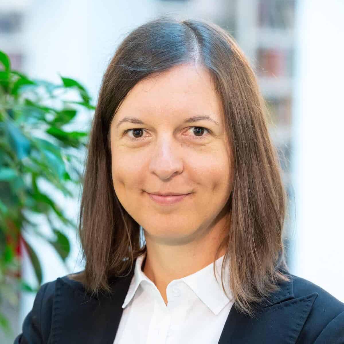 A woman with straight brown hair, wearing a white shirt and black blazer, stands with arms folded and smiles slightly. A green plant and blurred background are visible behind her.