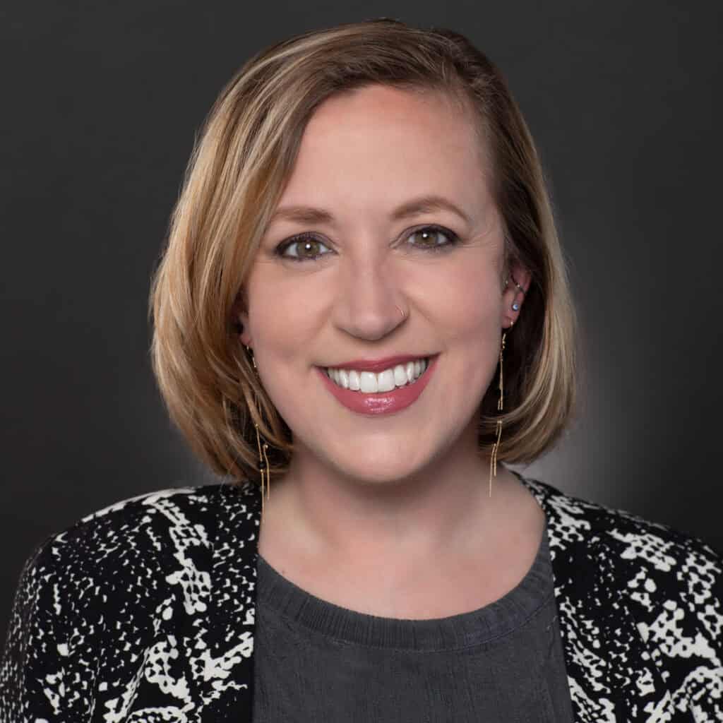 A woman with blonde bobbed hair smiles at the camera. She is wearing a patterned black and white jacket, a grey top, and gold dangling earrings, with a dark, plain background behind her.