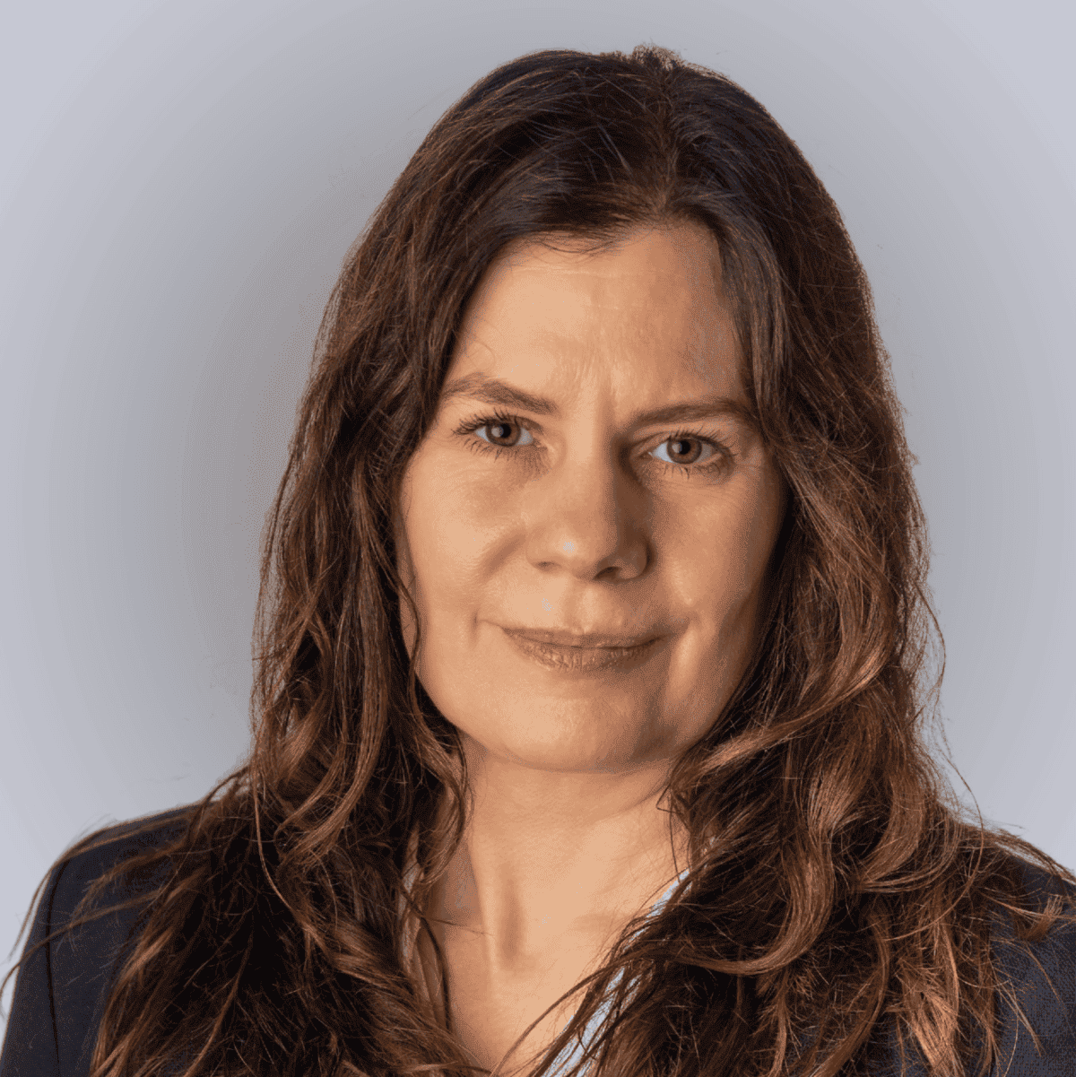 A woman with long brown hair wearing a navy blazer and a white blouse with small patterns, posing in front of a plain light grey background.