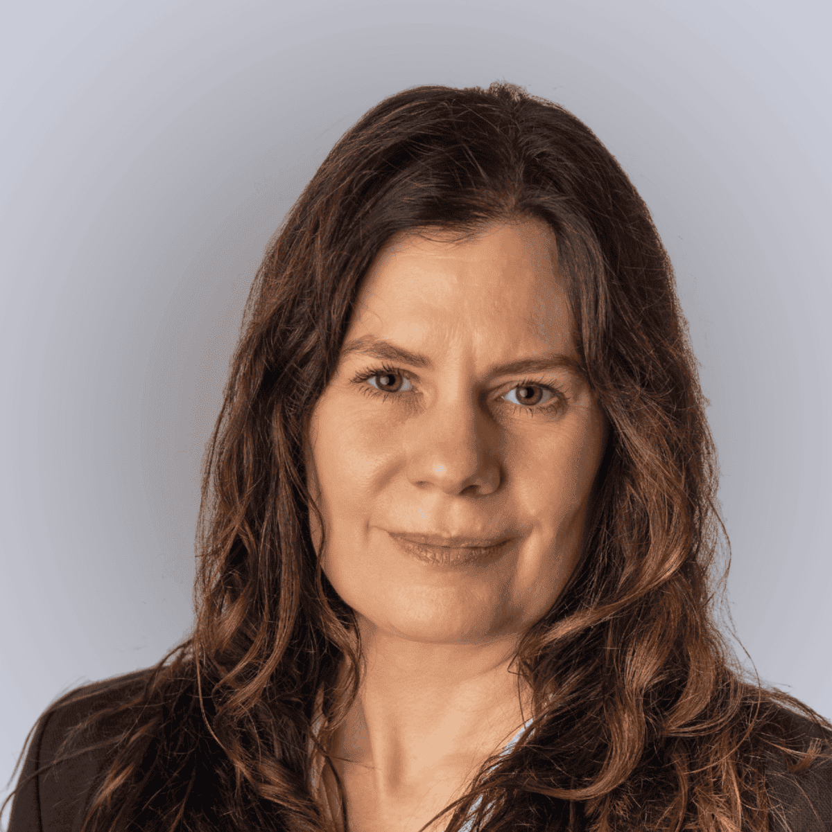 A woman with long brown hair wearing a navy blazer and a white blouse with small patterns, posing in front of a plain light grey background.