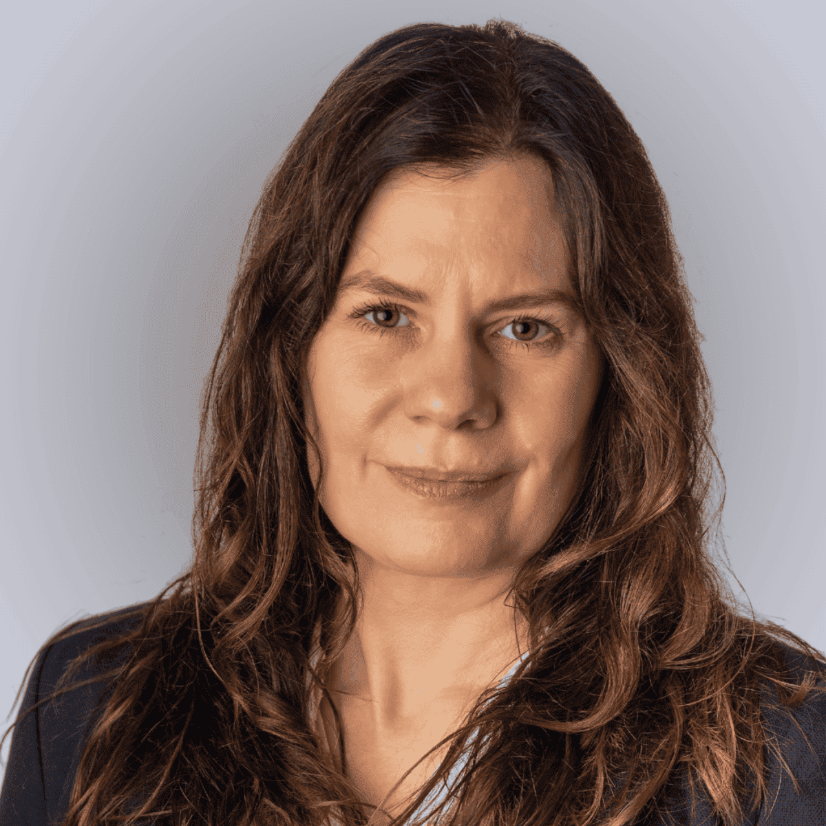 A woman with long brown hair wearing a navy blazer and a white blouse with small patterns, posing in front of a plain light grey background.